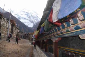 Trekkers passing a painted mani wall with prayer flags and snow peak backdrop on the Annapurna Circuit Trek