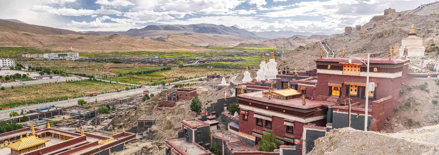 Aerial view of a Tibetan monastery complex with maroon and white buildings and golden stupas, set against a valley town and dry mountain ranges under a cloudy sky.